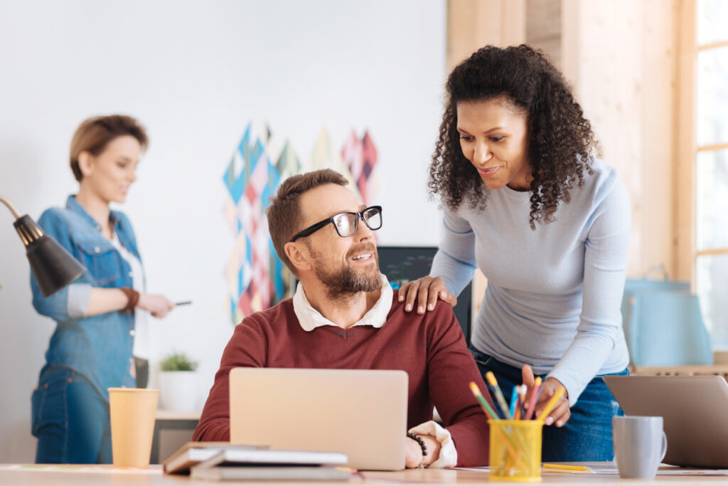 Handsome content bearded man working on his laptop while sitting at the table with smiling black woman standing behind him offering support